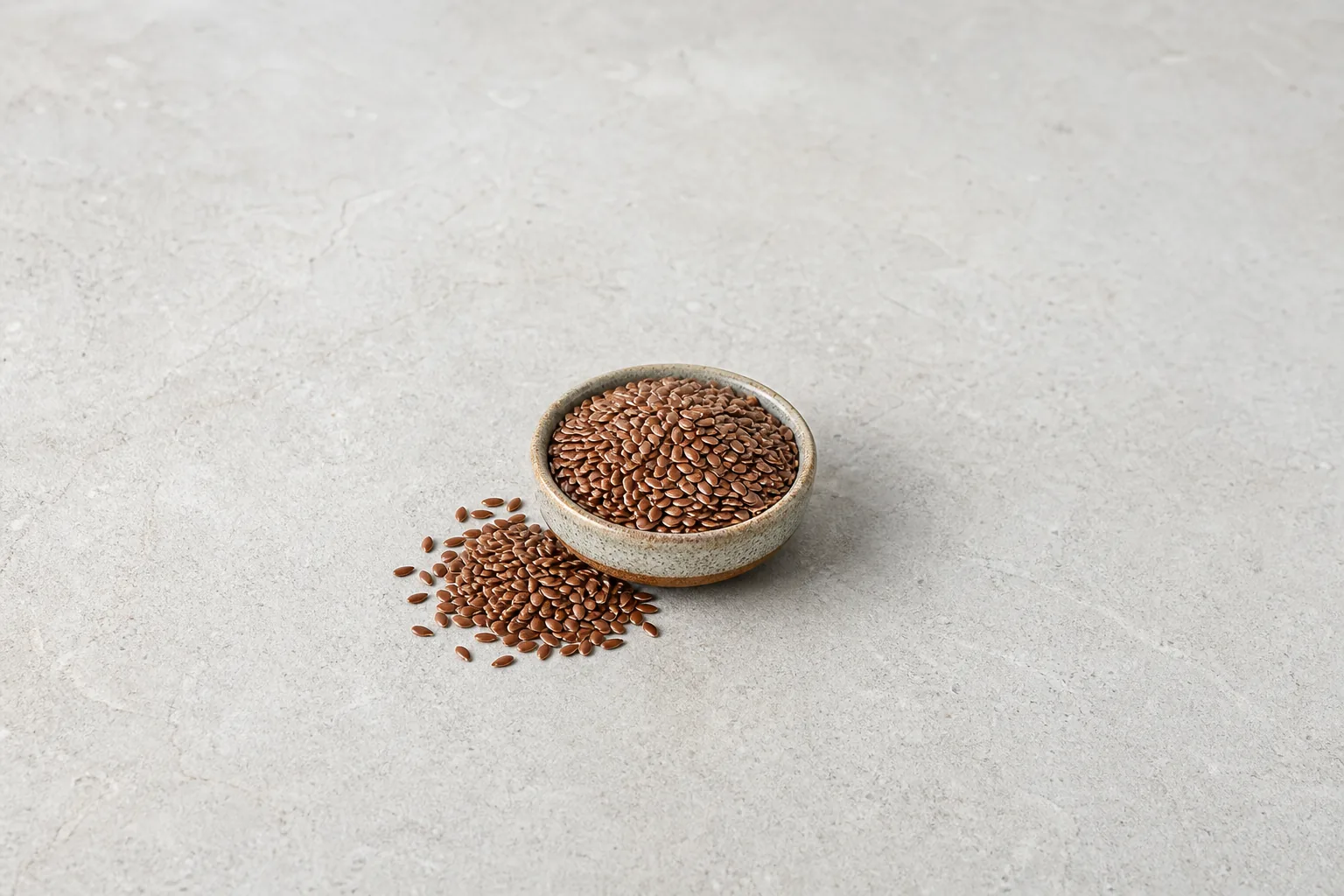 A small bowl of glossy brown, flattened flaxseeds with several loose seeds on pale stone.