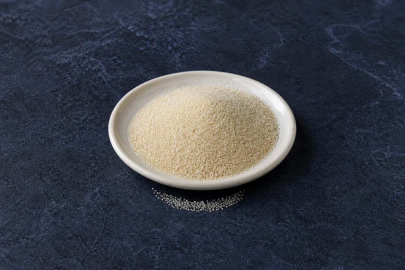 A shallow cream saucer of tiny pale dry fonio grains with a short spill.