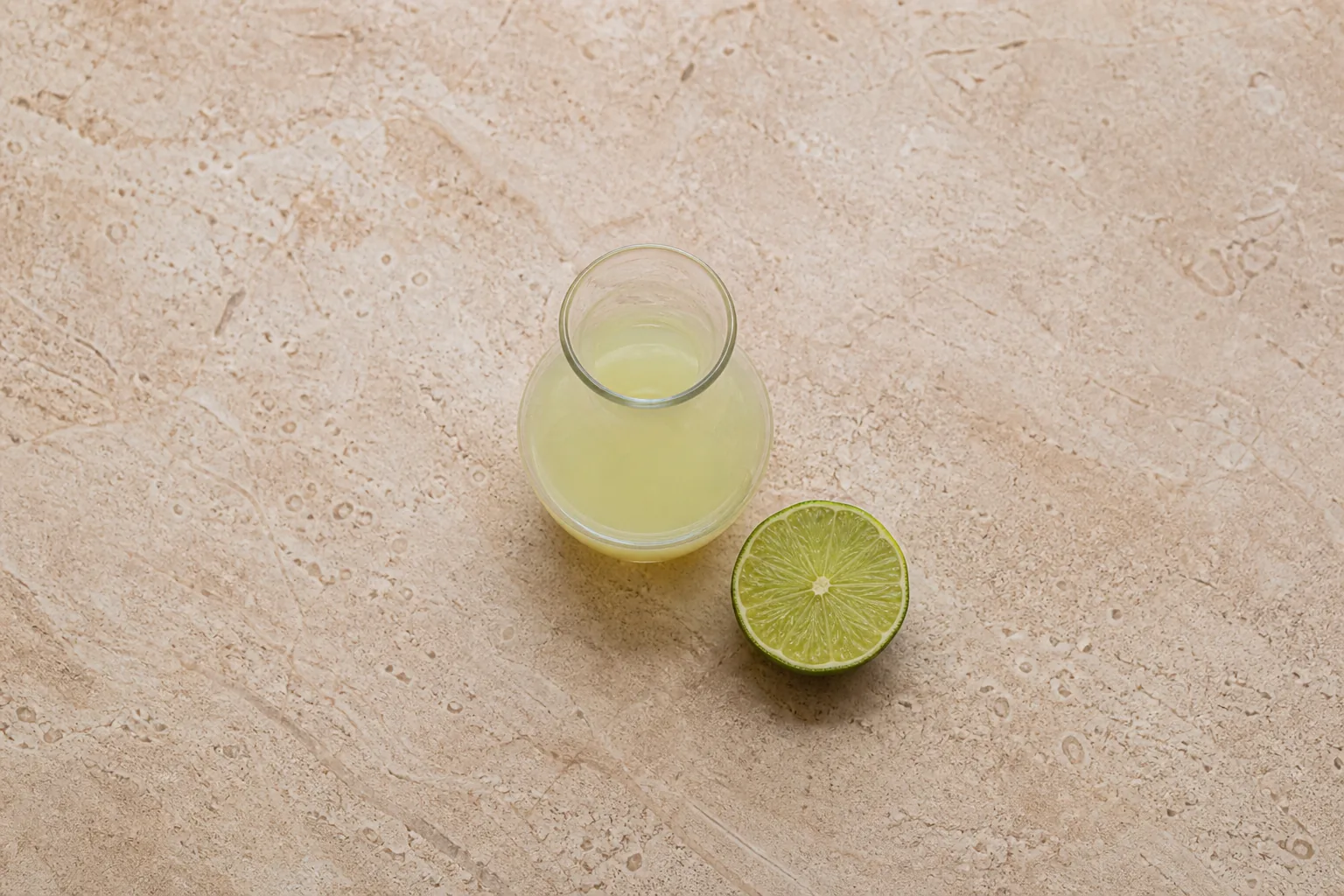 A small clear carafe of pale greenish-yellow juice beside one cut green lime half on warm stone.