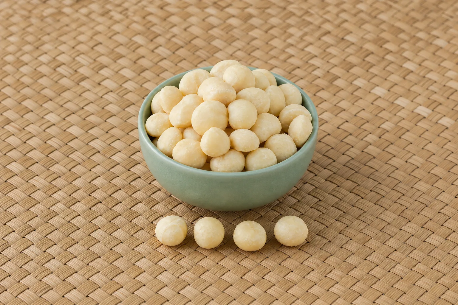 A small bowl of pale raw shelled macadamia nuts with several whole kernels beside it.