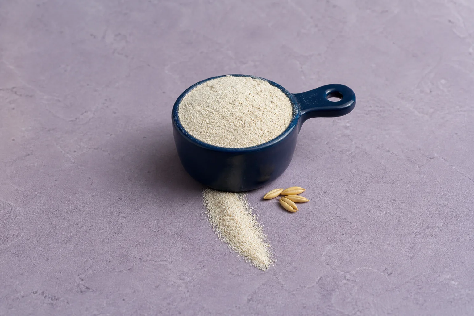 A navy measuring cup of fine pale beige flour with a short flour trail and several oat groats.