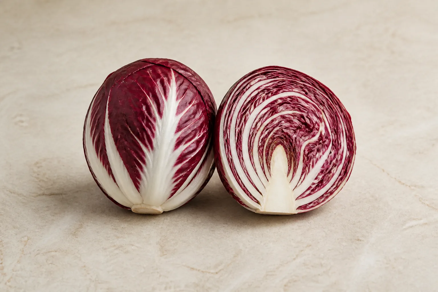 A raw burgundy radicchio head and one cut half showing tightly layered maroon leaves with bold white ribs.