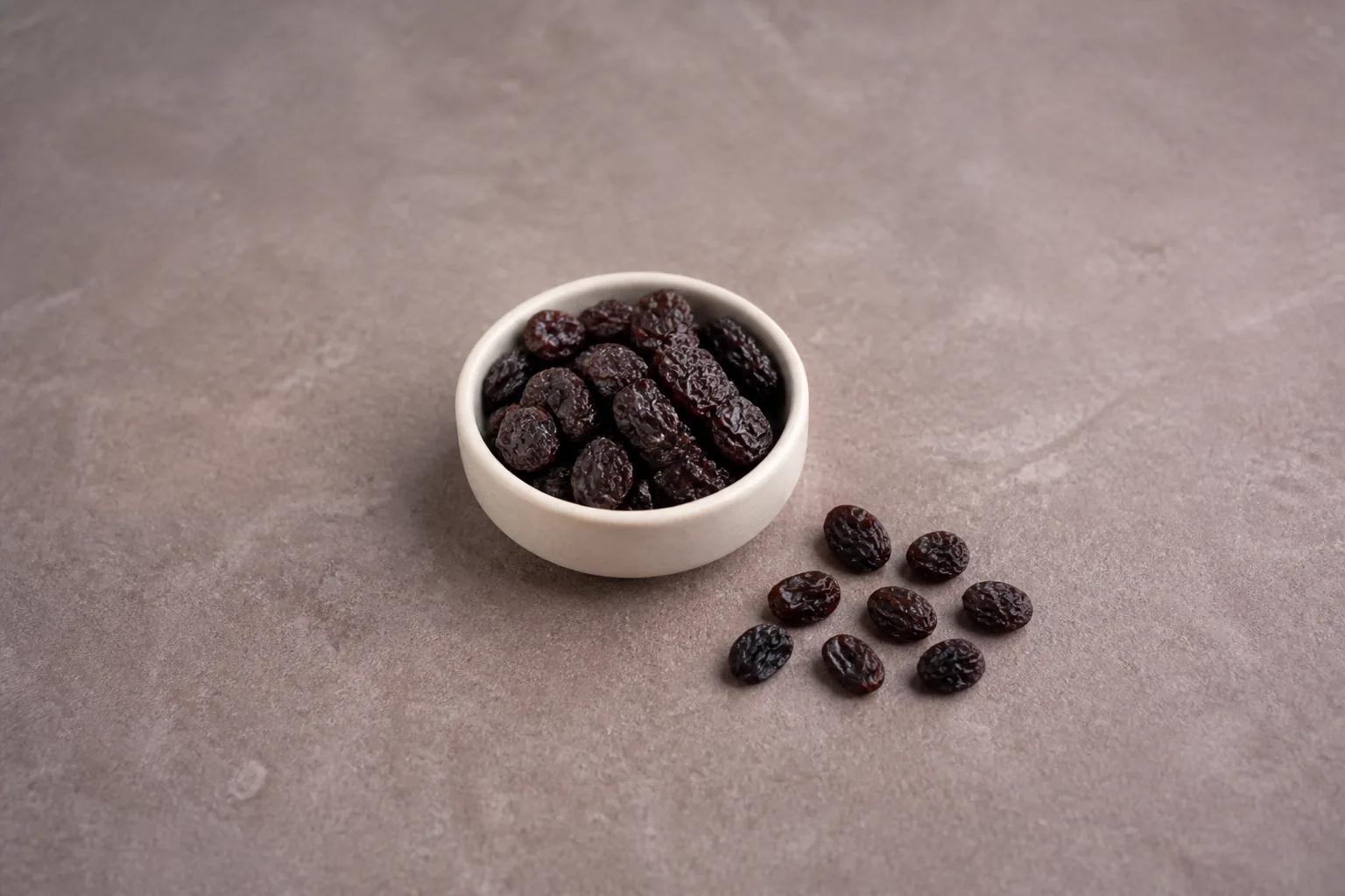 Dark seedless raisins in a shallow bowl with a compact group of loose raisins nearby.