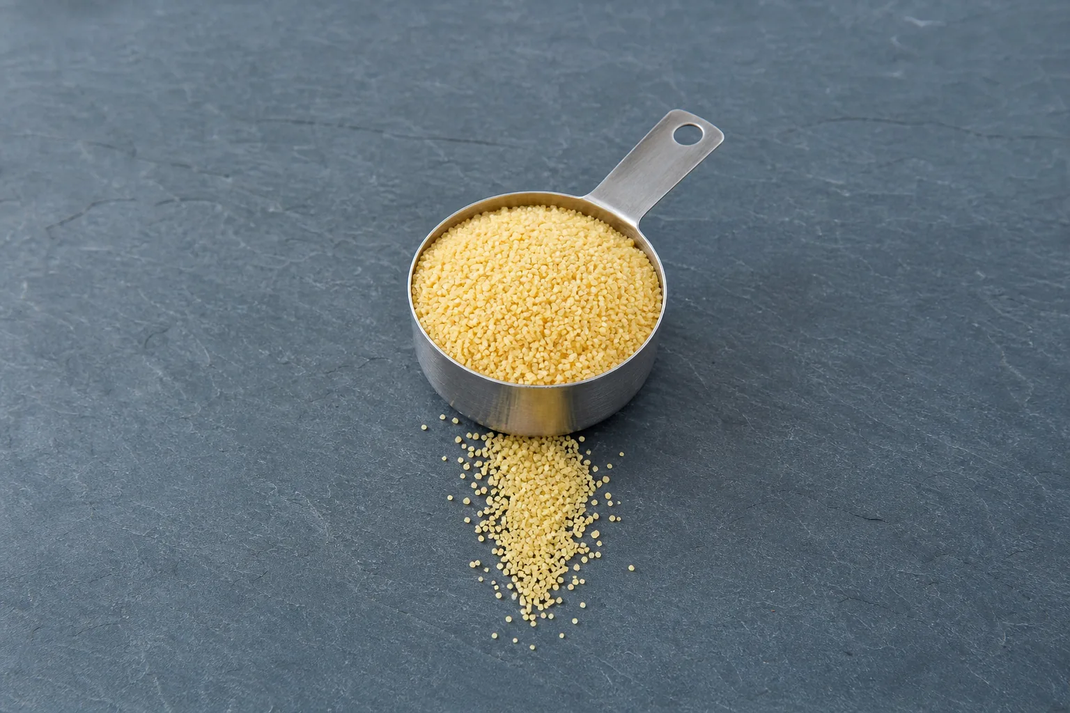 A metal scoop of coarse pale golden dry semolina with a short trail of granules.