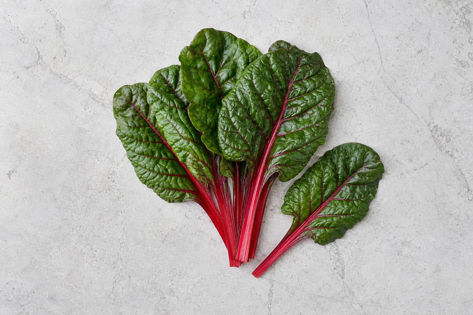 Raw Swiss-chard leaves with broad green blades and thick red stems on pale stone.